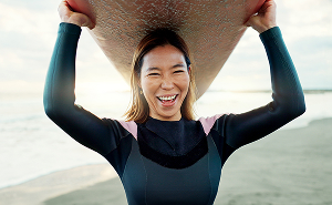 Smiling woman holding a surfboard at the beach, symbolising vitality and confidence from hormone therapy.
