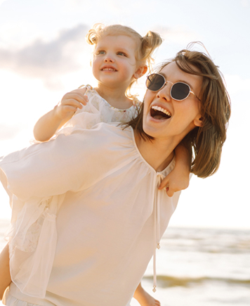 Happy woman carrying her young daughter outdoors in sunlight, symbolizing energy, vitality, and awareness of early signs of premenopause.
