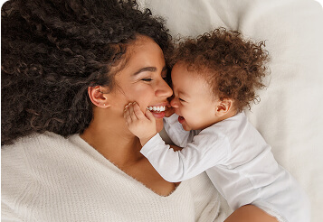 Smiling mother lying on a bed, holding and laughing with her baby, representing restored fertility and hormone balance after PCOS treatment.