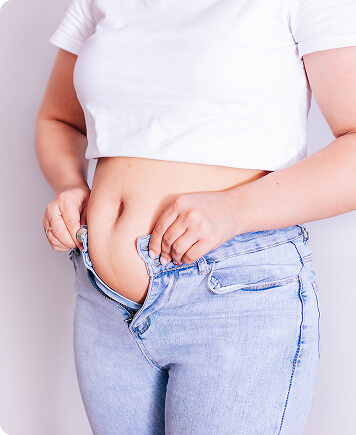 Woman standing and unbuttoning her jeans, highlighting bloating and abdominal weight gain related to cortisol and hormonal imbalance.
