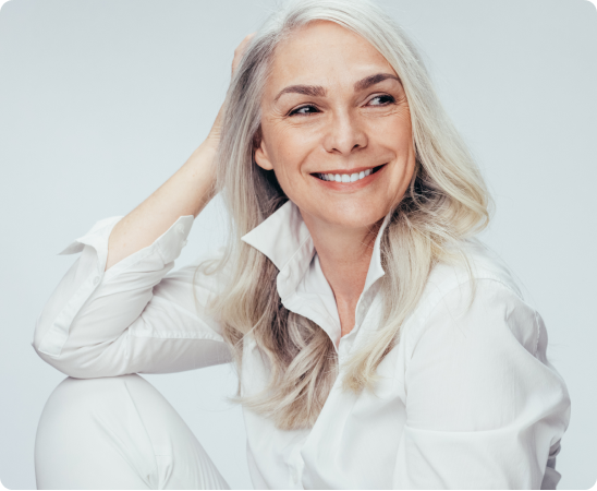 Smiling woman with long grey hair wearing a white shirt, sitting relaxed against a light background.