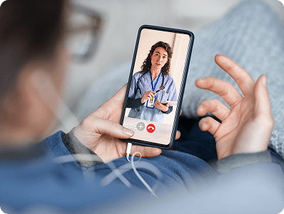 A person has a telehealth video call on their smartphone with a female doctor in blue scrubs.