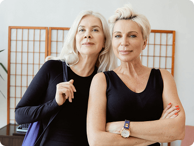 Two mature women with blonde hair wearing black tops pose together in front of a wooden room divider.