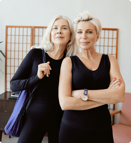 Two mature women with blonde hair wearing black tops pose together in front of a wooden room divider.