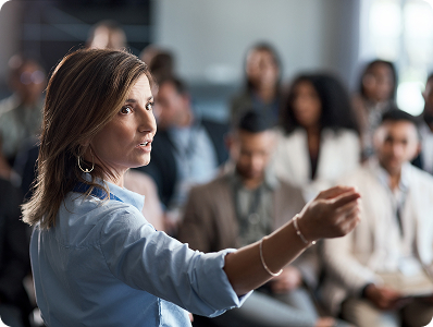 A woman in a blue shirt gestures while speaking to an audience in a professional setting.