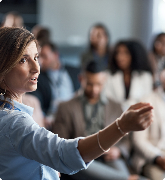 A woman in a blue shirt gestures while speaking to an audience in a professional setting.