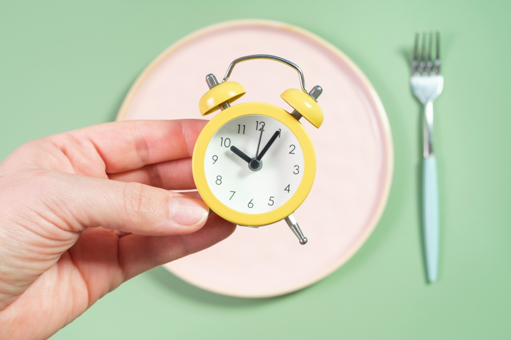 Female hand holding alarm clock against empty plate intermittent fasting concept