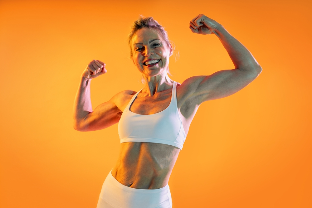 happy mature woman in sportswear flexing her arms in front of orange background