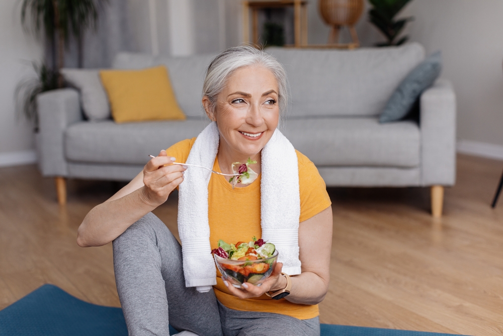Happy senior woman eating fresh vegetable salad sitting on yoga mat after home workout